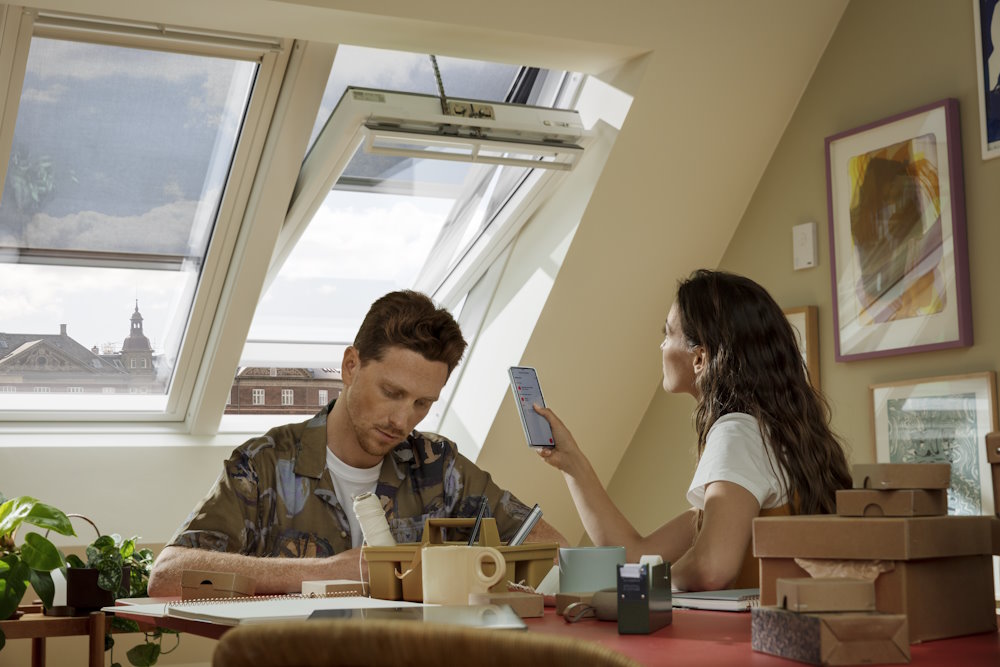 Zwei Personen sitzen unter einem geöffneten Dachfenster, eine Person hält ein Smartphone hoch.